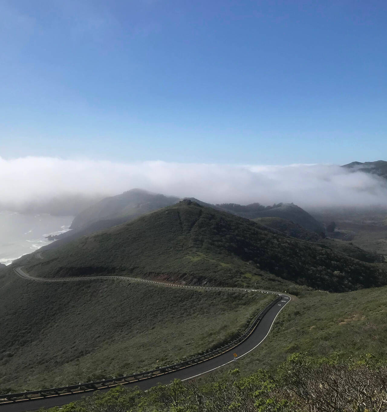 A road snakes across a hillside as marine fog encroaches on the coast behind