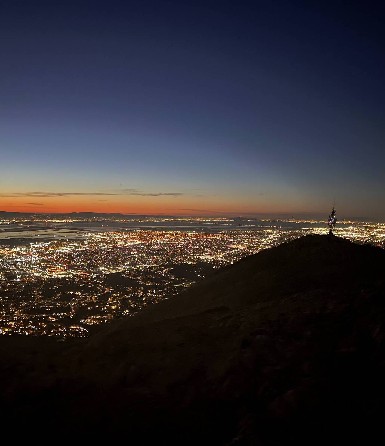 Looking over the top of Mission Peak, the sun glows orange and blue as the lights begin to illuminate the Bay Area