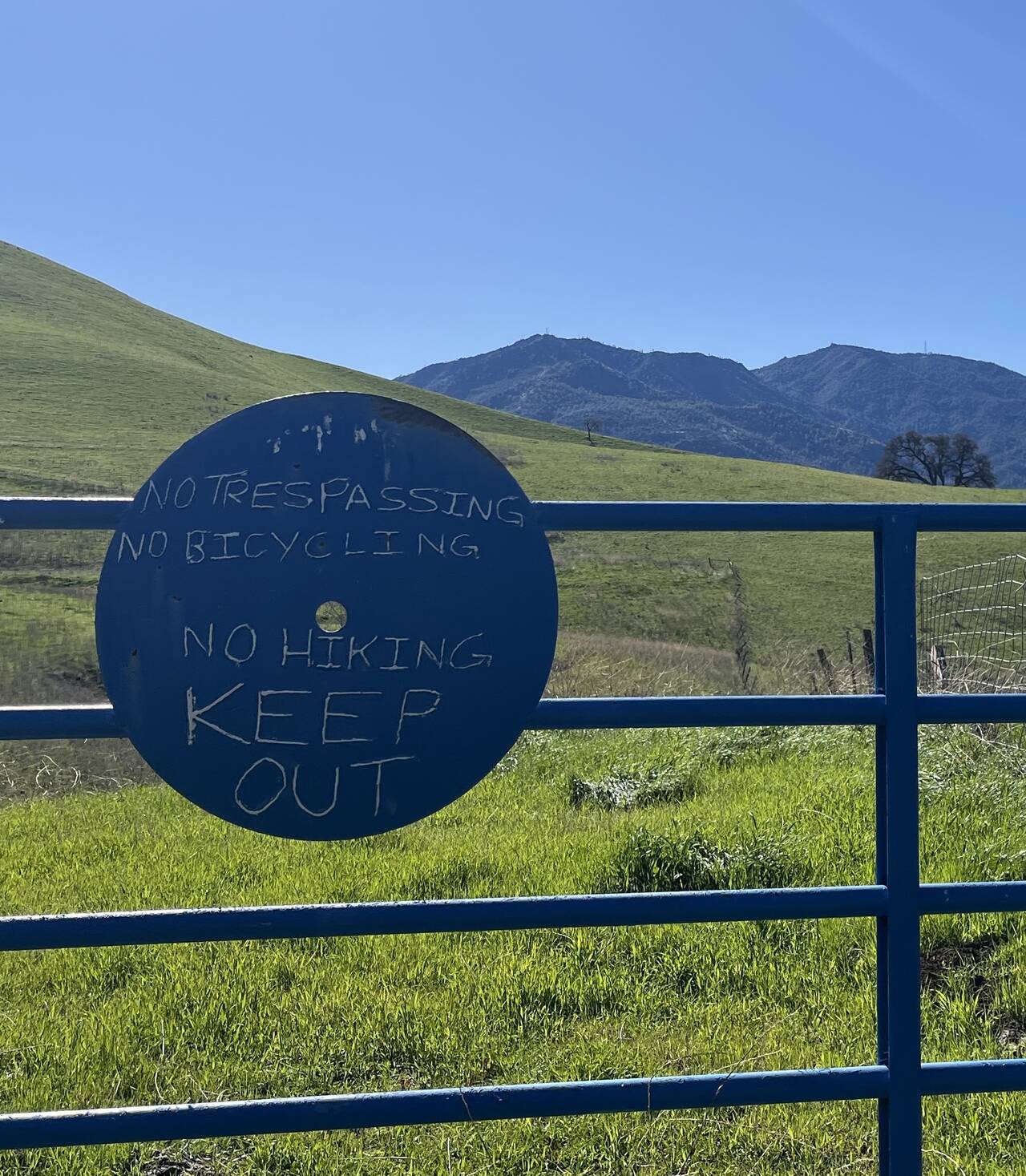 A sign on a blue gate reads "No tresspassing; no bicycling; no hiking; KEEP OUT." Beyond that, green fields and Mount Diablo.