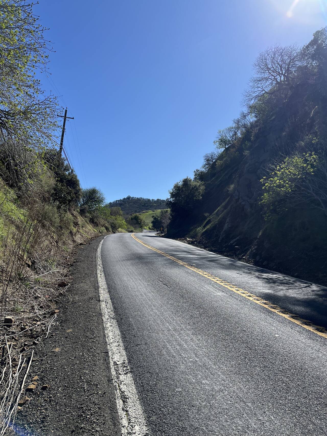 A country highway with a narrow shoulder. Terrain slopes steeply upward on both sides of the road.