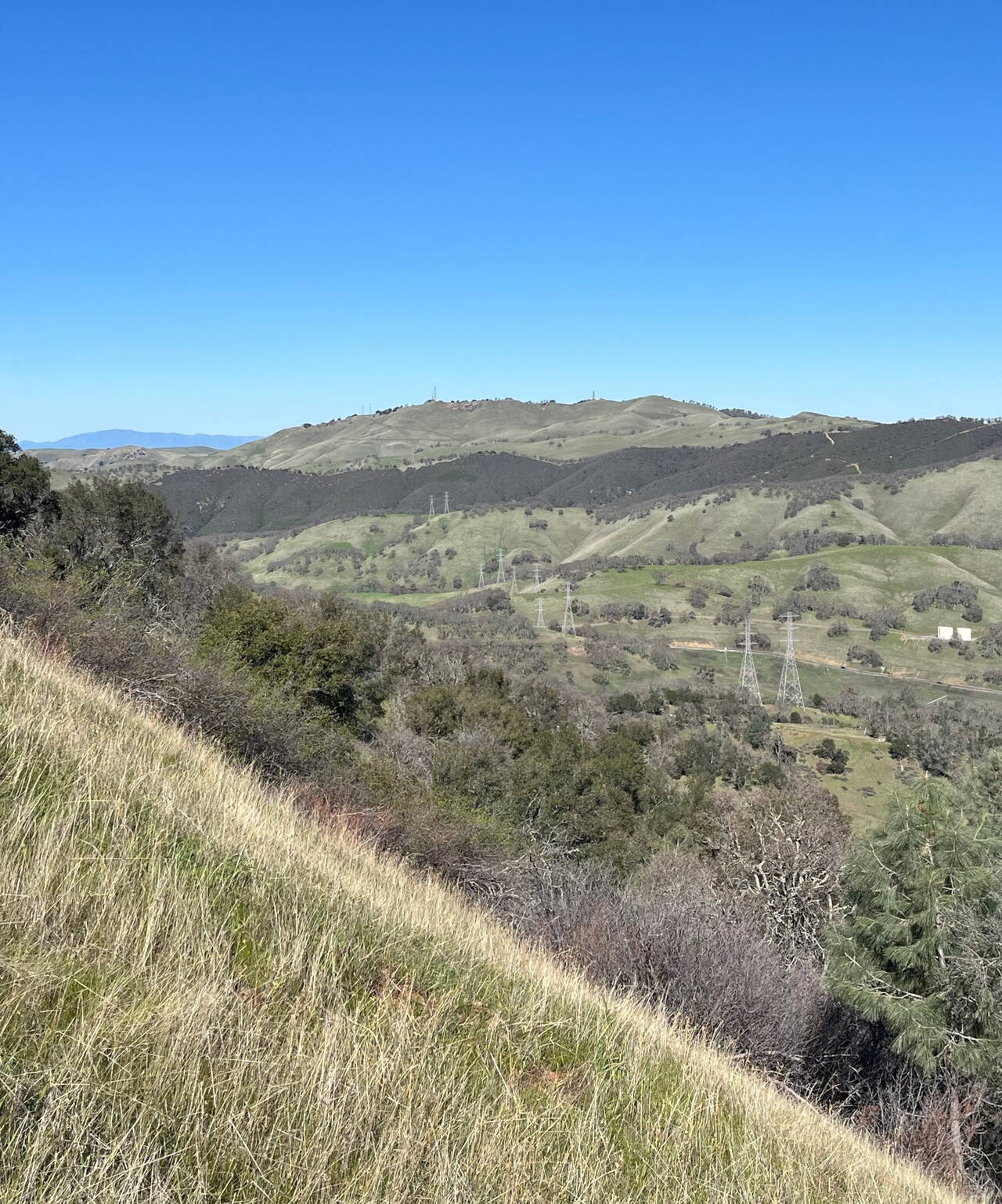 A north-facing photograph taken from the trail. Hills extend into the distance, and a side-view of a grassy hillside shows the slope of the trail.