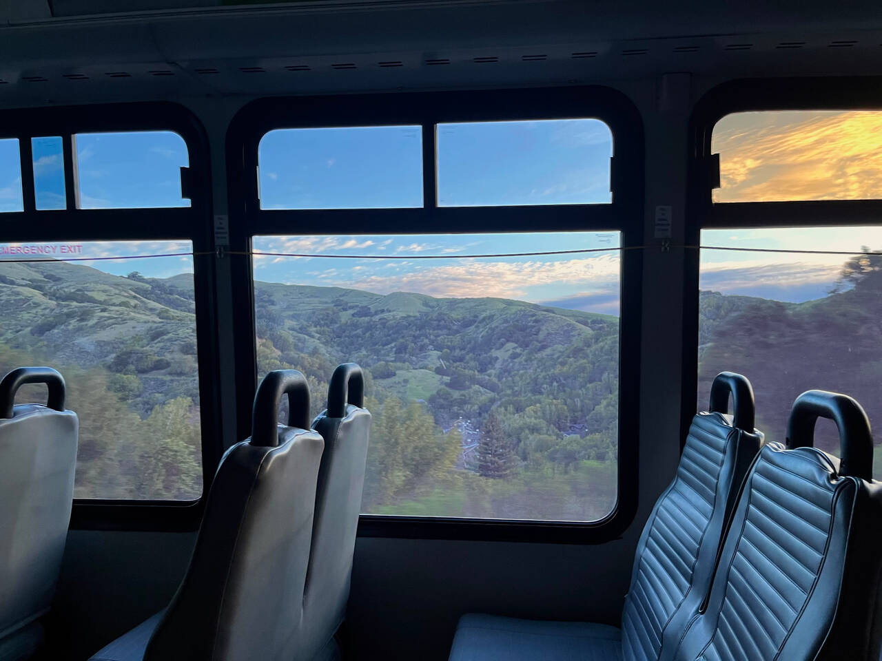 A picture looking sideways, out the window of the bus. Seats are visible in the foreground. Hills and a sunrise are visible out the window.