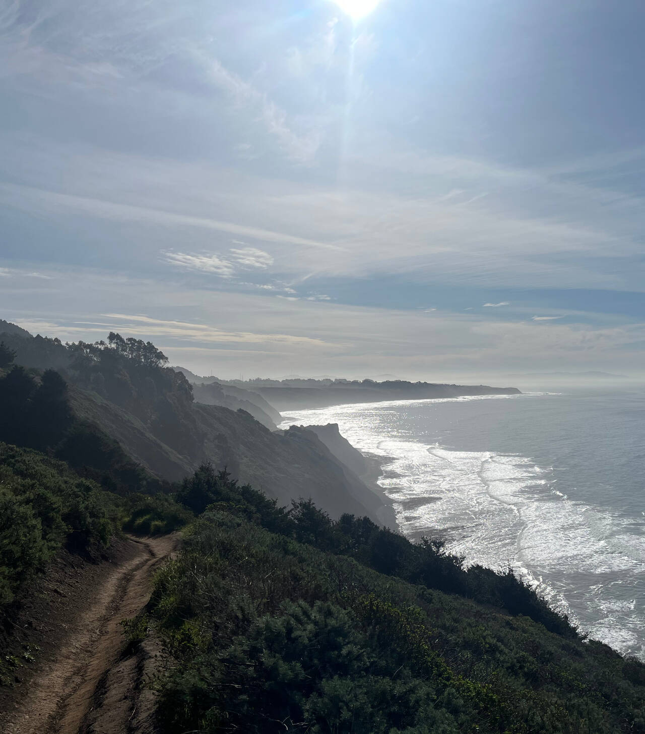 Looking south along the ocean, waves crash onto the shore below. The air is hazy.